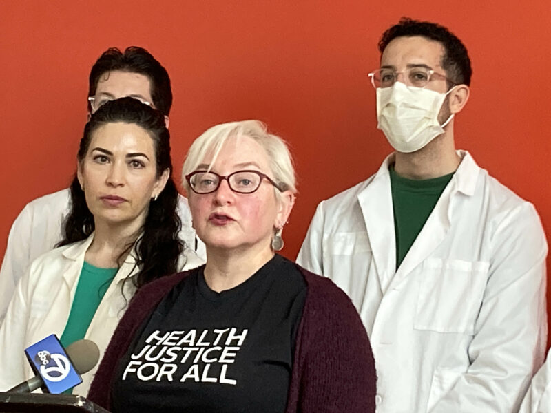 A woman with short, platinum blonde hair wearing glasses and a T-shirt printed with the words "Heath Justice for All" speaks into a microphone at a podium where she is standing near a woman and two men, all wearing white lab coats, in front of a red wall.