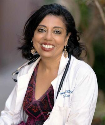 A smiling woman with dark hair wears a white lab coat with a stethoscope draped around her neck. 