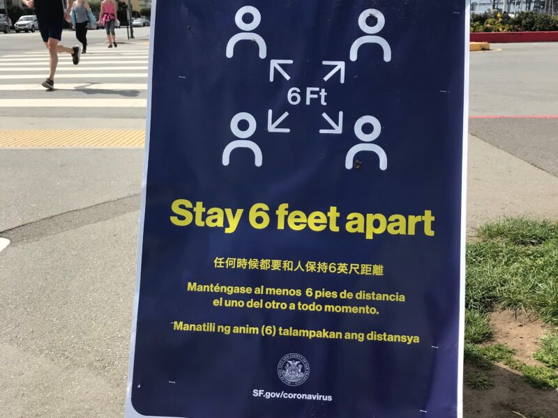 A poster with a blue background and white and yellow graphics and lettering placed near a sidewalk urges people to "Stay 6 feet apart."