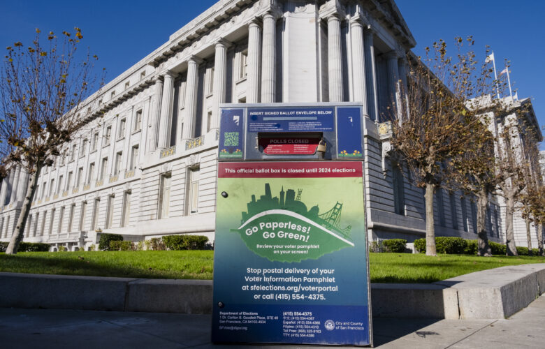 A metal ballot box covered with colorful decals featuring election information is located on a sidewalk in front of a green lawn with the tall columns of San Francisco's City Hall in the background.