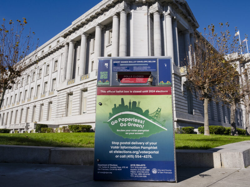 A metal ballot box covered with colorful decals featuring election information is located on a sidewalk in front of a green lawn with the tall columns of San Francisco's City Hall in the background.