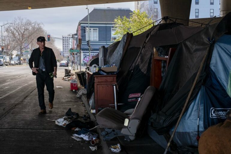 A man wearing dark clothing and a baseball cap walks under a freeway overpass and past a tent and other makeshift structures surrounded by small furniture and other household items.