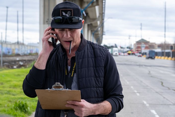 A man holding a clipboard and wearing a navy baseball cap holds a cell phone to his ear while standing near a roadway.