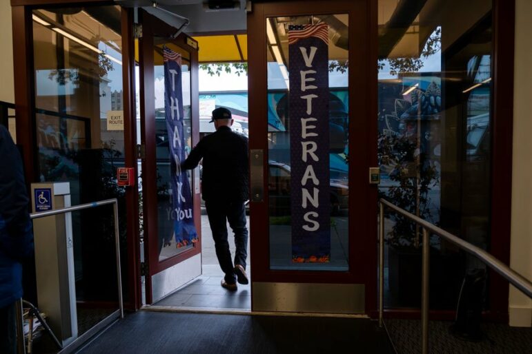 A man exits a building through a pair of glass doors with red trim bearing banners that read "thank you" and "veterans."
