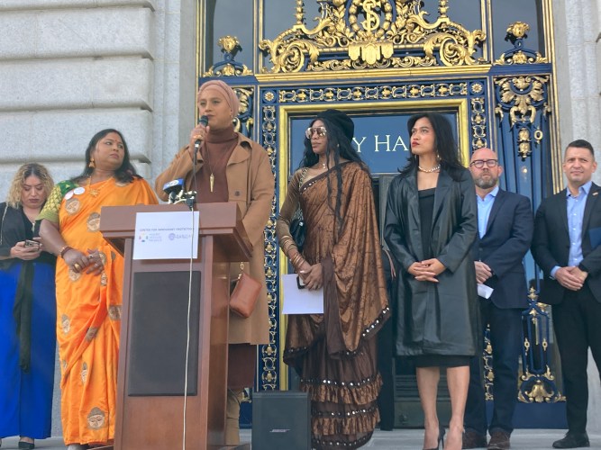 A woman at a lectern holding a microphone stands with several other people on the steps of San Francisco City Hall.