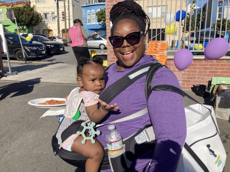 A woman wearing sunglasses and a purple top smiles broadly while holding an infant child in a carrier that she wears.