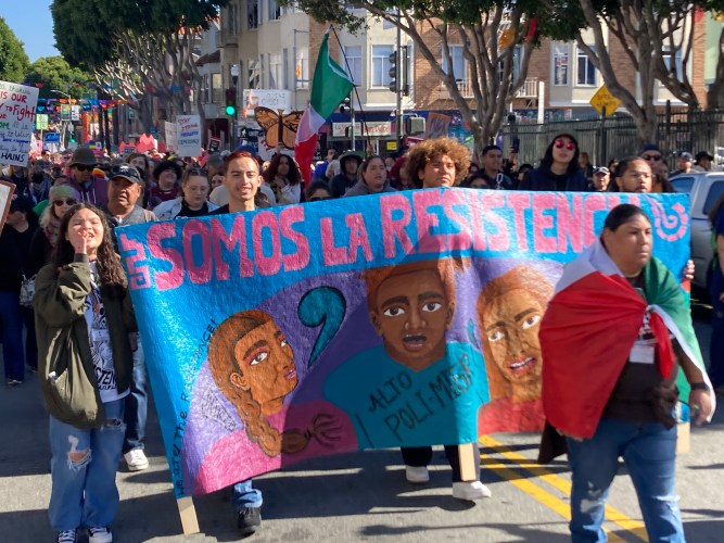 Throngs of people carrying signs and flags walk down an urban street behind four people holding a large blue banner reading "Somos La Resistencia" in Spanish in large pink letters, with a smaller English translation reading "We are the resistance."