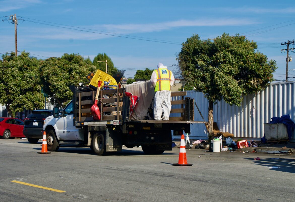 A person in a neon vest stands on a truck bed that is filled with belongings, and to its right, items are scattered on the sidewalk.
