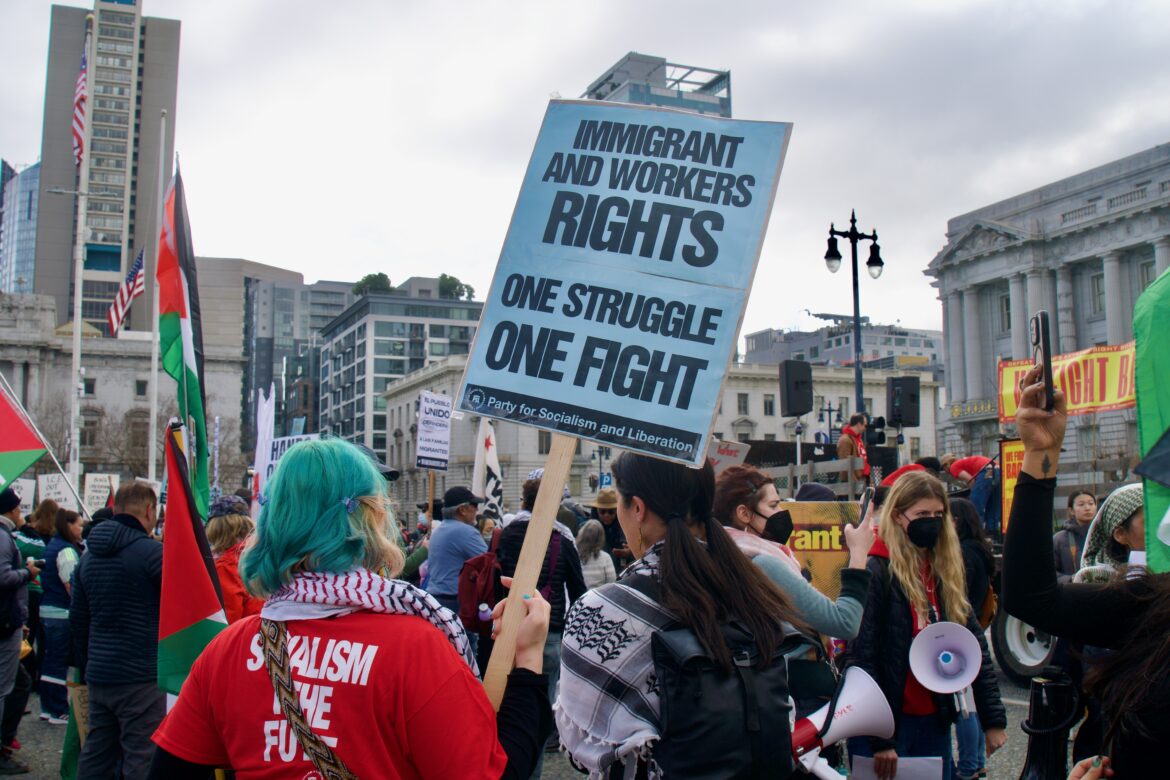In a crowd, a person with teal hair in a red shirt holds up a poster that says "IMMIGRANT AND WORKERS RIGHTS ONE STRUGGLE ONE FIGHT"