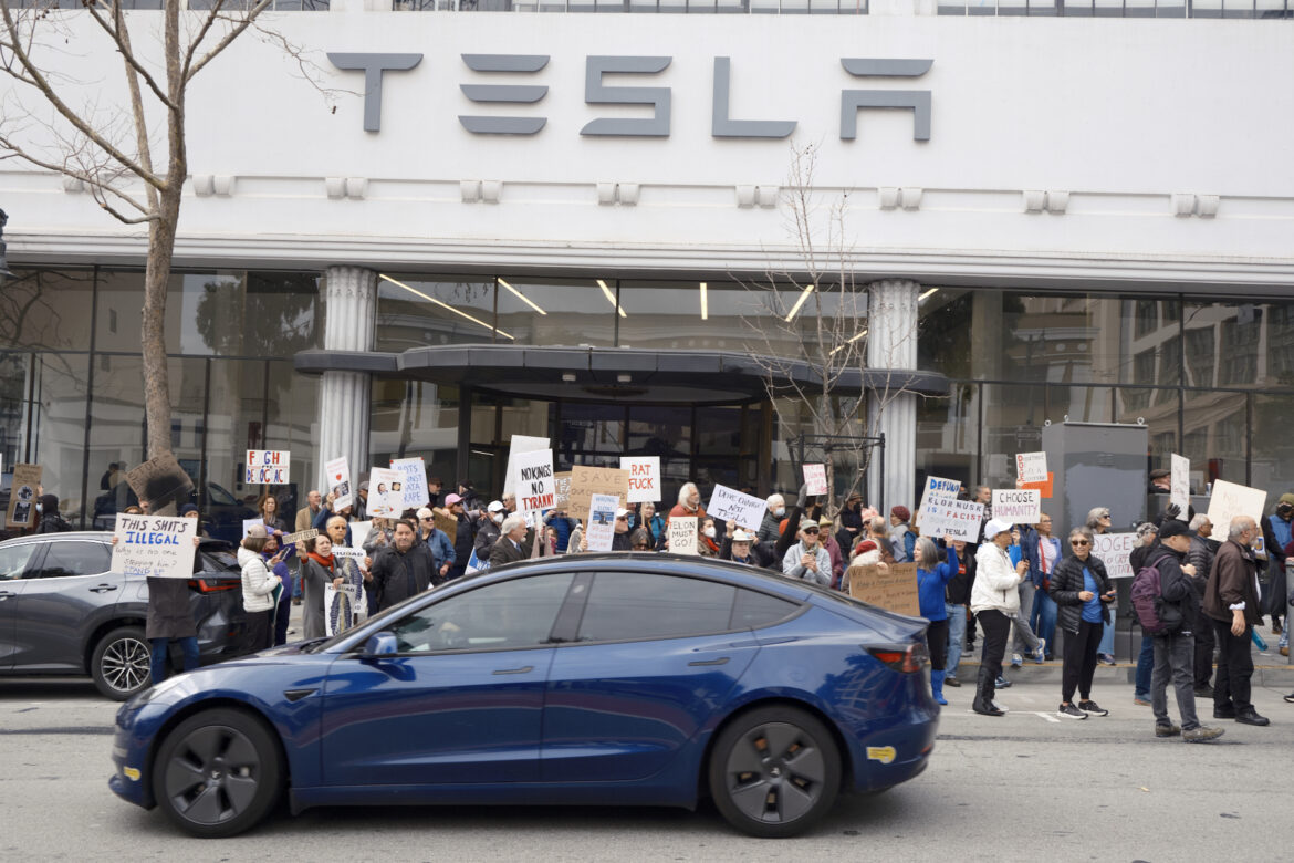 A blue Tesla drives past a crowd of protesters carrying signs in front of a light gray building with large plate glass windows and the word TESLA in stylized letters on its facade.