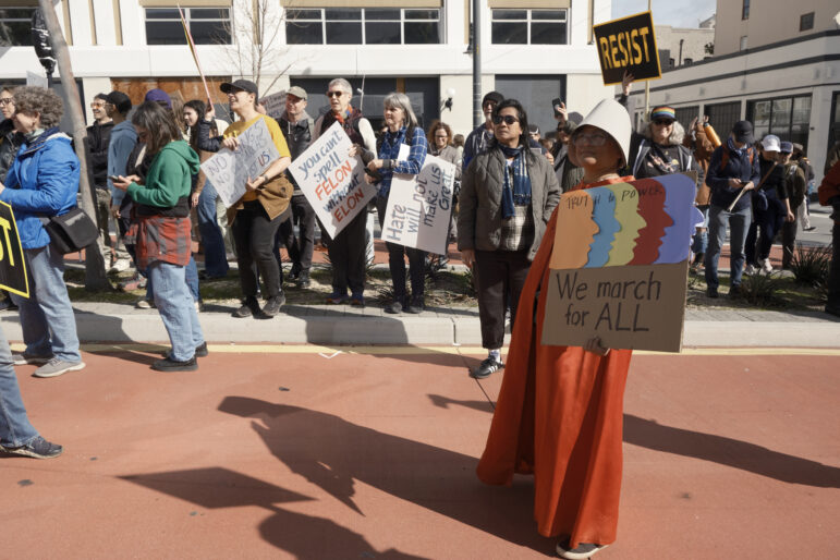 Protesters carrying signs stand on a sidewalk and spill into the street, standing in a red bus-rapid-transit lane, where a woman wearing a long red robe and a white pilgrim-style hat holds a colorful sign with two slogans: "truth to power" and "we march for all."