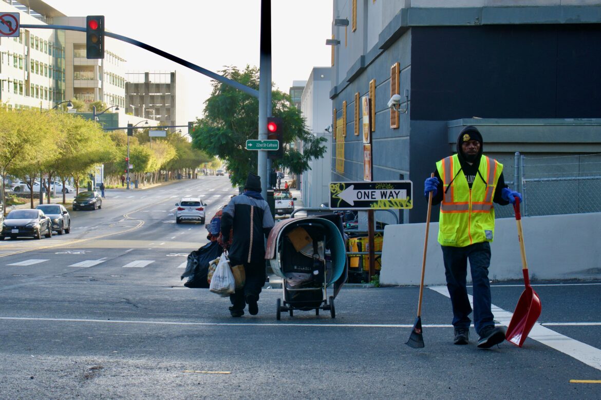A man in a neon yellow vest carries a shovel and rake across the street. In the background, another man is pulling a stroller filled with belongings to the sidewalk.