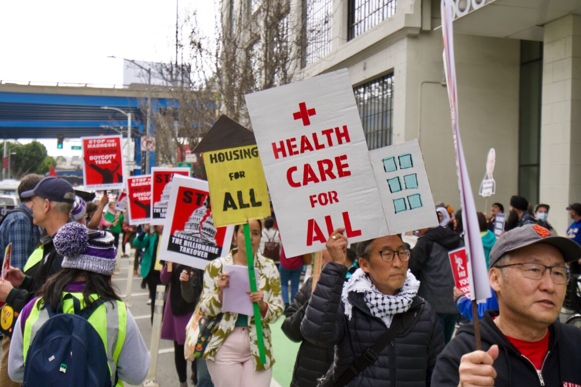 Protesters in bright clothing hold up colorful signs that say "HEALTHCARE FOR ALL," "HOUSING FOR ALL," AND "STOP THE BILLIONAIRE TAKEOVER."