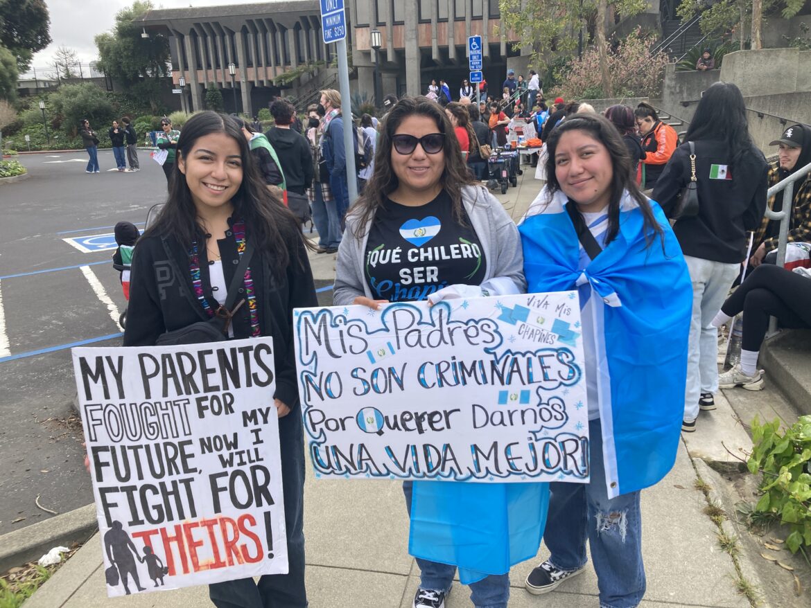 Three women smile and hold hand drawn signs reading "my parents fought for my future — now I will fight for theirs" and "mis padres no son criminales, por querer darnos una vida mejor."