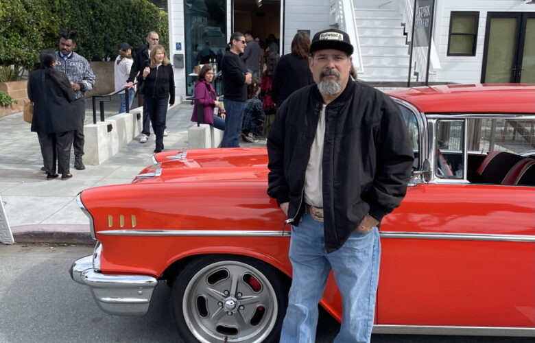 Artist Oscar Lopez is wearing jeans, a black jacket and a black hat while standing in front of a bright red classic car parked in front of a white clapboard building with "M Stark Gallery" in large letters over the door and several people mingling in the art gallery's front courtyard.