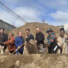 Nine people stand behind a mound of dirt, each holding a shovel in the ceremonial groundbreaking.