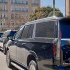 Several large black SUVs park in line along an urban street.
