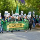 A group of dozens of people carrying signs march behind a green and yellow banner that says "AFSCME 3299"