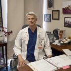 A man wearing blue scrubs and a white doctor's coat sits behind a desk in an office.