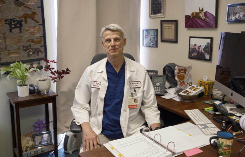 A man wearing blue scrubs and a white doctor's coat sits behind a desk in an office.