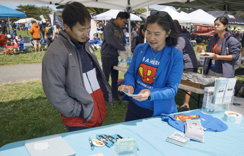 A woman wearing a bright blue jacket over a bright blue T-shirt with a red and yellow logo speaks with a man wearing a gray jacket over a shirt with broad blue, white and red stripes, as they stand by a table displaying printed materials under a shade structure at an outdoor community event.