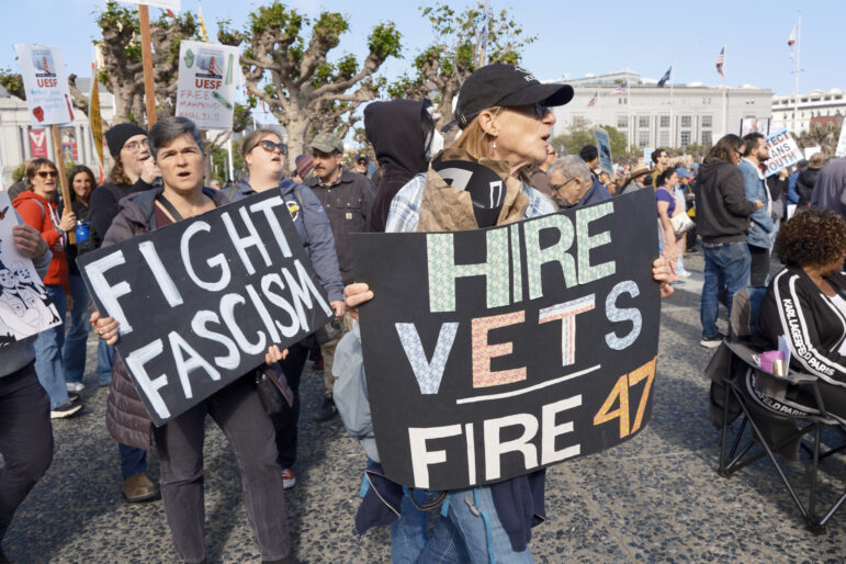 Protesters gathered outdoors carry signs, including ones with pastel letters on black poster boards reading "FIGHT FASCISM" and "HIRE VETS, FIRE 47."