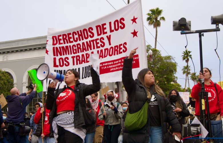 A woman surrounded by people yells into a bullhorn. Behind her, a white banner with red text reads "DERECHOS DE LOS IMMIGRANTES Y LOS TRABAJADORES"