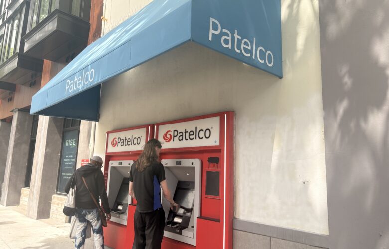 Two people use ATMs set into an exterior wall and surrounded by bright red frames capped by "Patelco" signs under a medium blue awning with Patelco in white lettering.