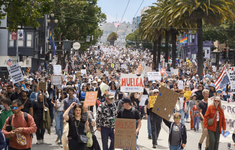 Thick crowds of protesters march down both sides of a boulevard, and a giant pink triangle is visible on the distant hillside behind them.