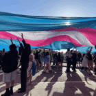Several people march holding a large white, pink and blue flag over their heads as others walk by in the background