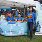 Two people sit behind a table and one stands in front of it. They are all wearing blue T-shires with a Superman-style logo featuring a "B" with a slogan: "B A Hero — Stop Liver Cancer."