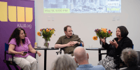 Three people sit in a discussion before an audience in front of a screen