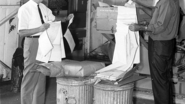 The Navy encouraged the disposal of paper records. In 1969, staff fed classified documents into industrial shredders in accordance with the service’s policy of “reducing paperwork to a minimum.” Source: National Archives and Records Administration. Two men holding large sheets of papers in front of a large shredding machine and trash cans in a black-and-white photo dated 1969 and labeled classified material