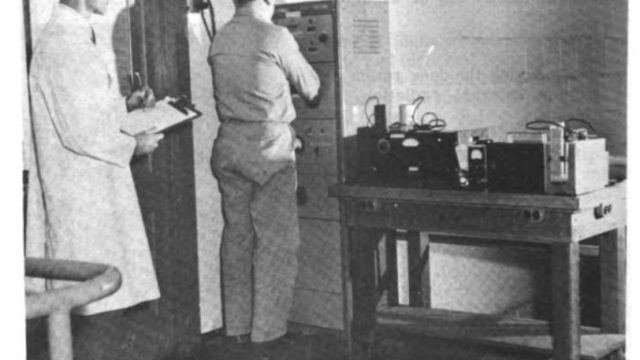In the 1950s, radiation scientists around the country worked with detectors that were imprecise and often underestimated exposure. This image shows measurements of radioactivity on exposed personnel at the Naval Research Laboratory in Monterey, Calif. Source: U.S. Navy. grainy black-and-white photo of two men in a laboratory, one standing in front of a large machine and another standing behind him in a white lab coat and holding a clipboard, with a caption saying
