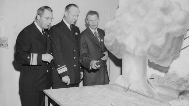 As scientific director of the Naval Radiological Defense Laboratory, Paul C. Tompkins, far right, shared military officials’ interest in ways troops might survive a nuclear war. With him are naval research chief Frederick R. Furth and Bureau of Ships chief Albert G. Mumma. Source: San Francisco Examiner Archive at UC Berkeley. Three men in suits, two of them with military adornments, pose before a diorama of a mushroom cloud and ships at sea in a black-and-white photo