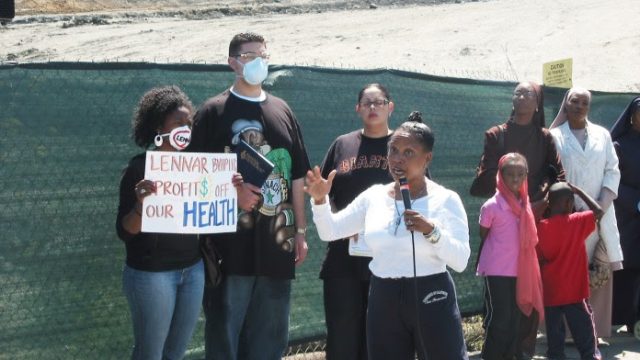 Ahimsa Porter Sumchai has supported neighborhood advocates for years in their quest for answers about pollution and community health in Bayview-Hunters Point. In 2007, she spoke in front of Parcel A, formerly part of a larger Superfund cleanup site. Ahimsa Porter Sumchai speaking to a crowd of people in front of a fence with a mound of soil in the background, with another woman holding a sign saying