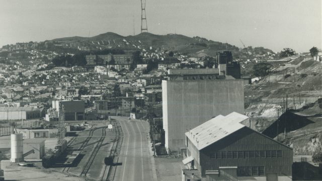 Recent research suggests contamination from the shipyard and radiation lab (white, nearly windowless building, center) has migrated into the nearby Bayview-Hunters Point neighborhood. Source: National Archives and Records Administration. black-and-white historical photo of shipyard buildings with neighborhoods and hills in the background