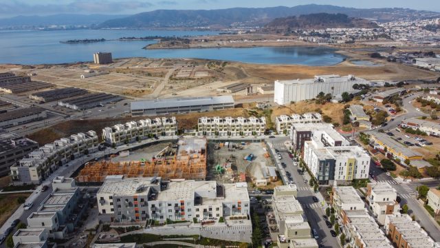 The threat of radioactive pollution — underground and in dust kicked up by construction — continues to be a concern for activists and residents near the former headquarters of the Navy’s radiation lab (white, nearly windowless building, upper right). aerial photo of dozens of housing units under construction in the foreground, and fields, industrial buildings and water in the background