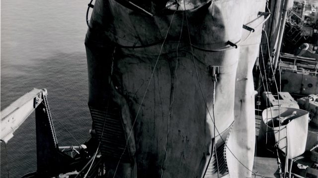 The Navy sent workers aboard the USS Crittenden, which was damaged in a U.S. nuclear weapons test in the Pacific, despite not knowing how much gas masks would help in filtering out airborne plutonium. Source: National Archives and Records Administration. part of the superstructure of a ship is dented and warped in a black-and-white historical photo