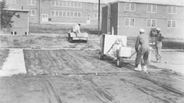 A 1965 lab report describes an experiment on methods to remove sod after a nuclear attack. Workers with little protective gear were exposed to radioactive simulated fallout while performing cleanup and measurement. Source: Naval Radiological Defense Laboratory report via Nuclear Testing Archive, Las Vegas. Men operate heavy equipment on a lawn in front of low-slung brick buildings in a black-and-white historical photo.