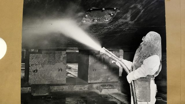 Cold War-era military officials knew radiation posed a “subtle, lurking hazard” to workers attempting to clean contaminated warships at the Hunters Point Naval Shipyard. Source: National Archives and Records Administration. A person in heavy protective gear draped over the head and torso, and wearing overalls, sprays water from a hose onto a part of a ship above in a black-and-white photo dated 1946