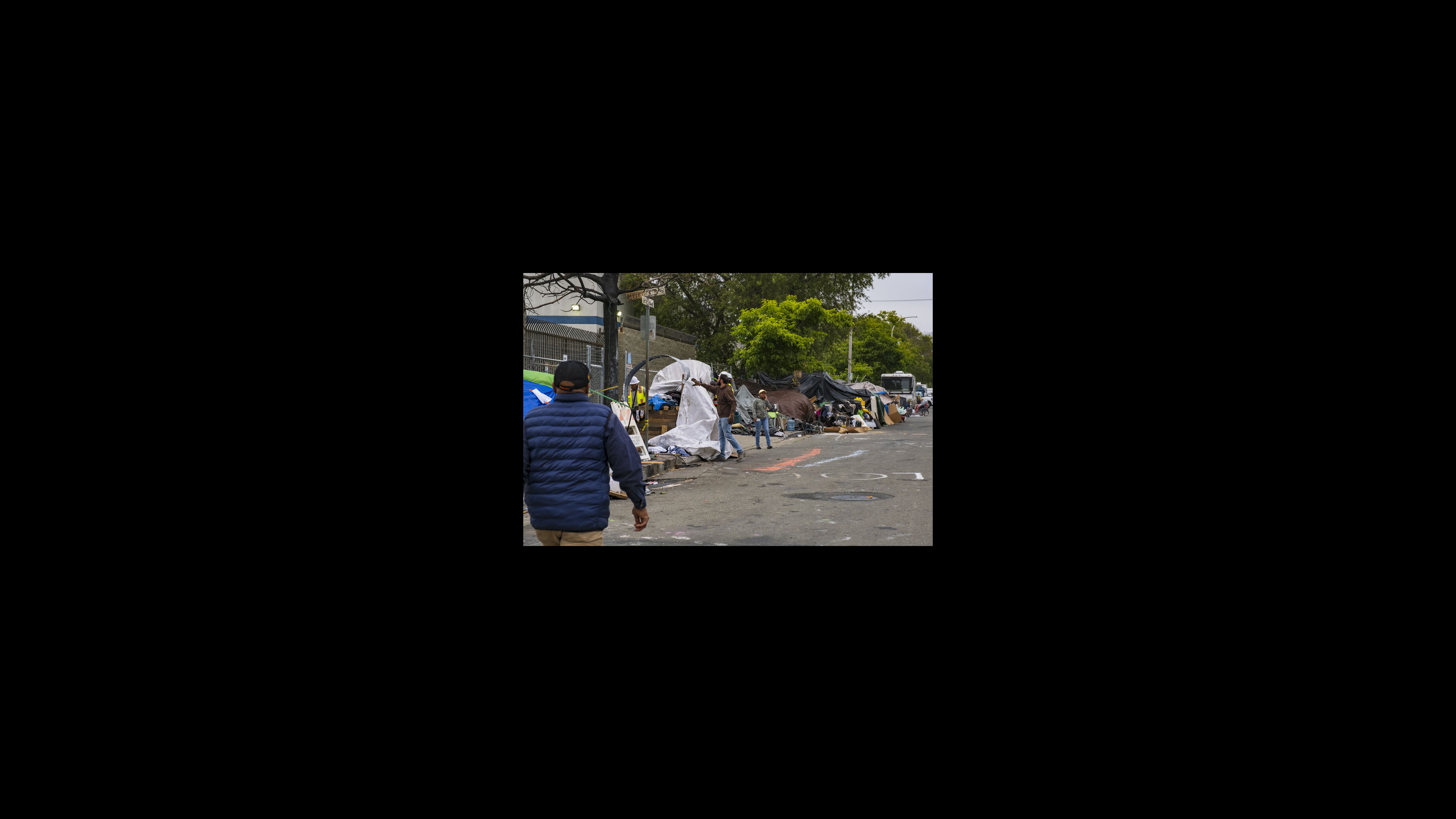The Berkeley Homeless Response Team prepares to close an encampment at Seventh and Harrison streets on May 23, 2023. Camp residents were asked to move down the street. Two accepted the city’s offer to put them up in rooms at the Berkeley Inn. Tents and RVs are lined up on one side of a street. City workers inspect a makeshift structure as they prepare to dismantle it.