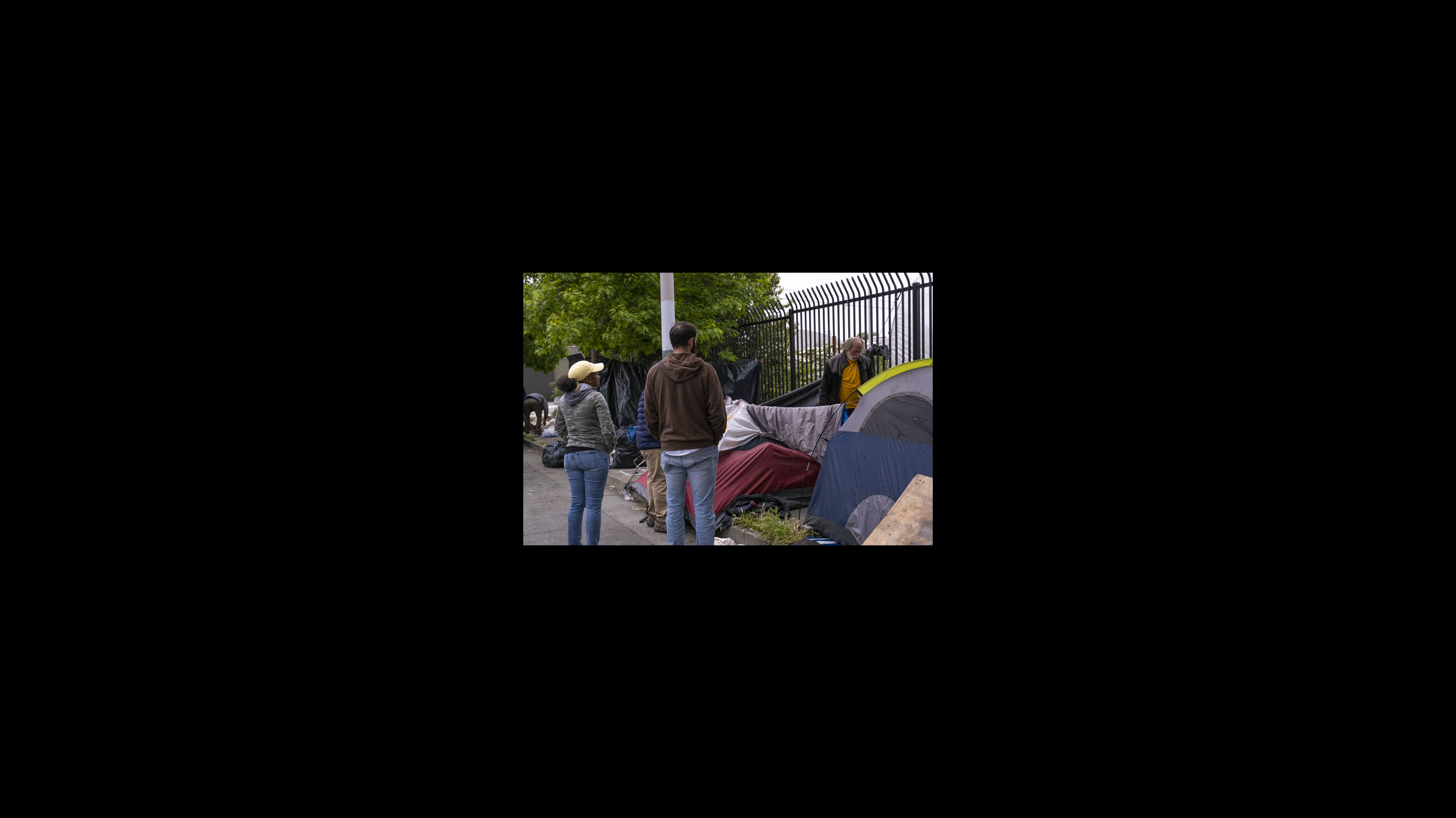 Peter Radu, assistant to the city manager, and Okeya Vance, supervisor of the Homeless Response Team, speak with Mike, who gives only his first name, about closing his section of the encampment and having him move into a hotel room. Mike relinquishes most of his property, which is scavenged by another neighbor and then thrown away. Two city workers stand on one side of a tent speaking to a man who is standing on the other side of it near a fence.