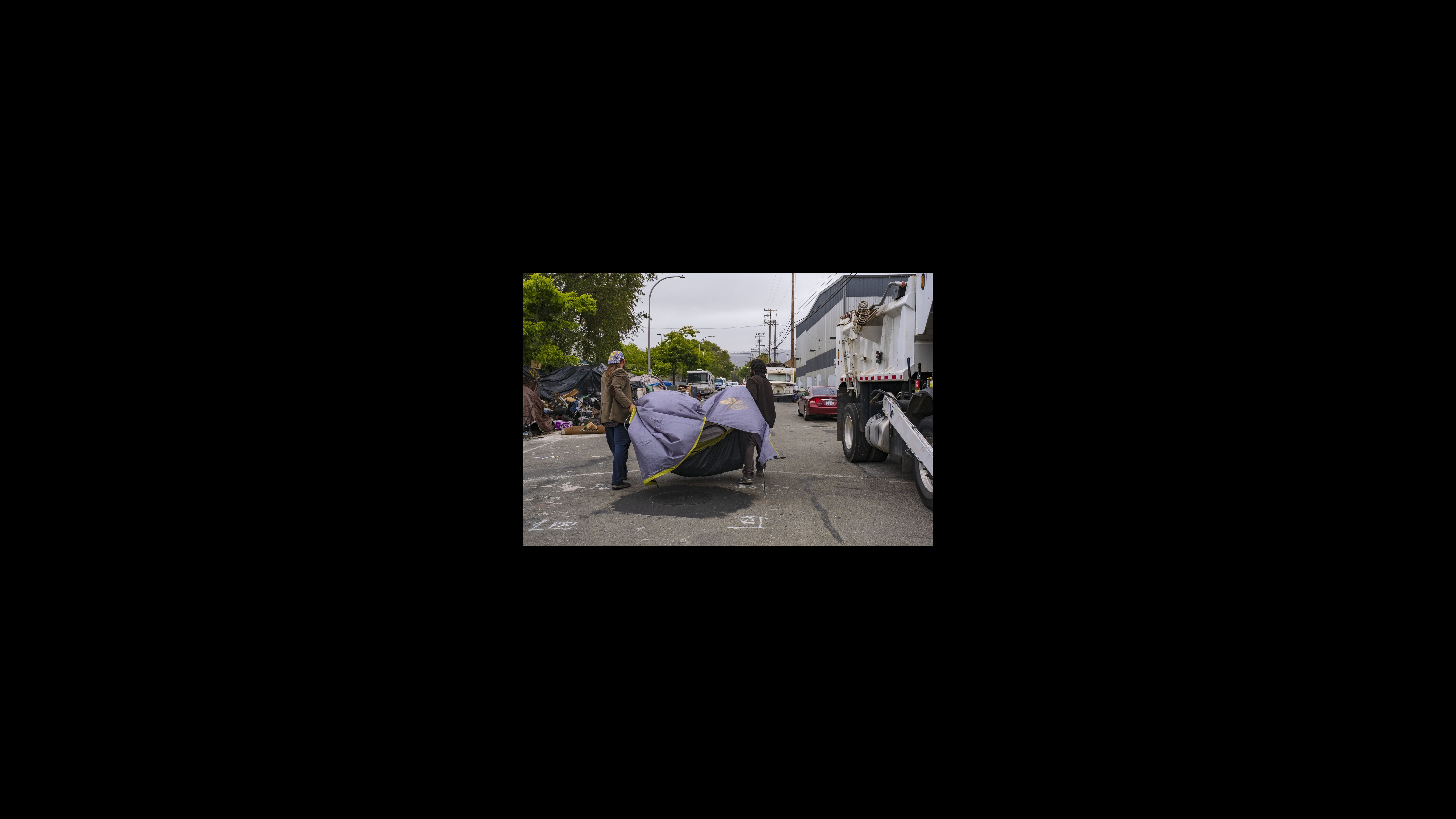 A.J., who goes by his first initials, receives help from an advocate, Gordon Gilmore, to carry his tent down the street. Although the encampment is not sanctioned, the city is allowing residents to camp temporarily on Eighth Street until more shelter beds become available. Two men with their backs facing the camera carry a gray tent down a street as they walk past a garbage truck and other tents and vehicles.