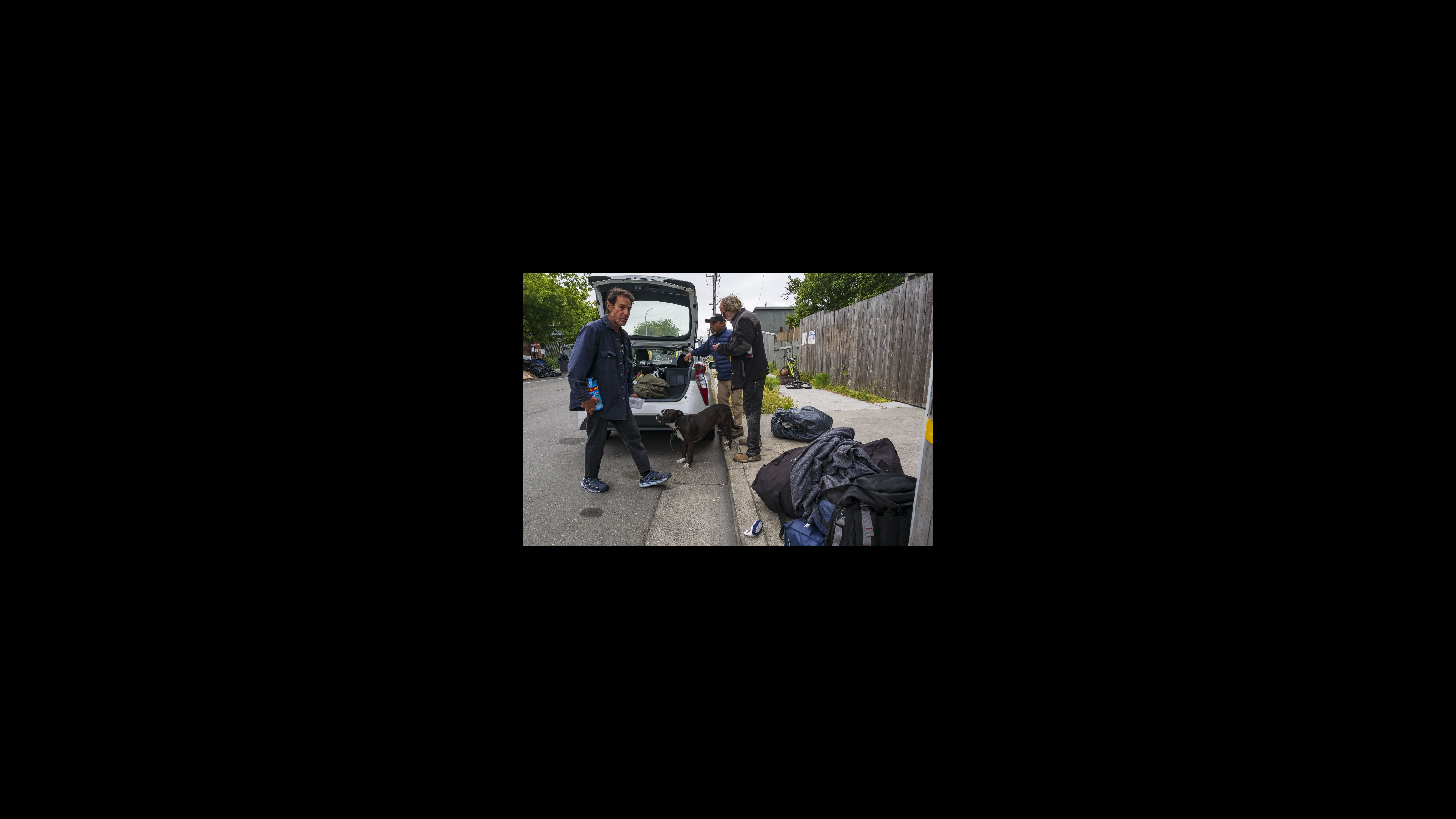 Tony Alcutt, a Homeless Response Team outreach worker (wearing a baseball cap), helps Chris, who walks behind the car, and Mike, who is standing on the sidewalk, load their personal items into the trunk of his car. He would drive them to the Berkeley Inn where the city is providing rooms. Three men and a brown and white dog stand near the back of a white car with its hatchback open. The men are moving bags and other items from the sidewalk to the car.