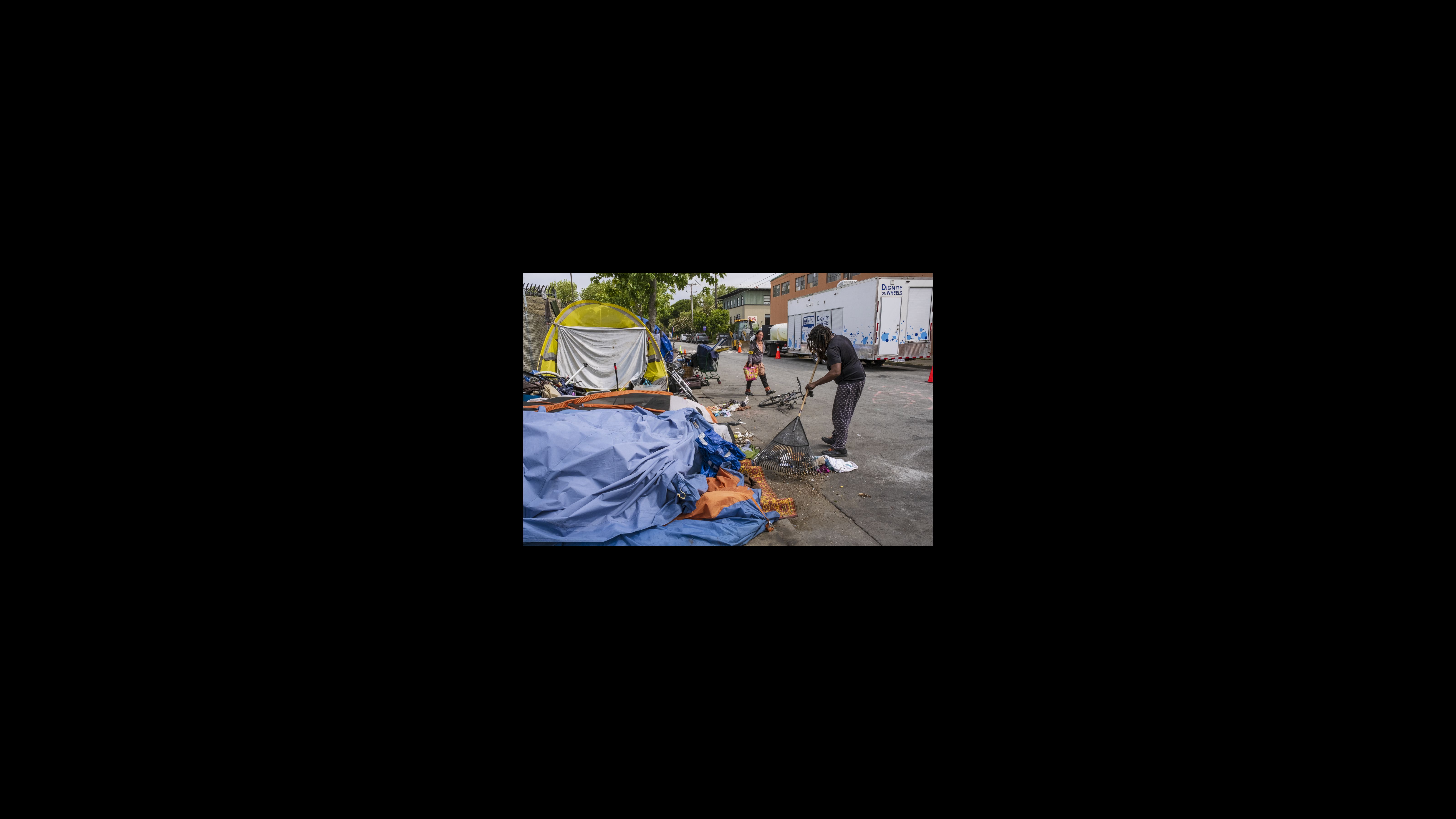 Mackie, who gives only his first name, rakes garbage around his tent into the road for Berkeley Public Works to pick up. Every Tuesday, a Dignity on Wheels truck comes to the neighborhood, providing 10-minute showers and two loads of laundry per person. While the resources are free, some residents shy away because they are required to fill out paperwork to access the showers and laundry services. A man rakes debris and other small trash items from around a tent into the street. Other tents stand nearby. A woman carrying a bag walks by in the background.