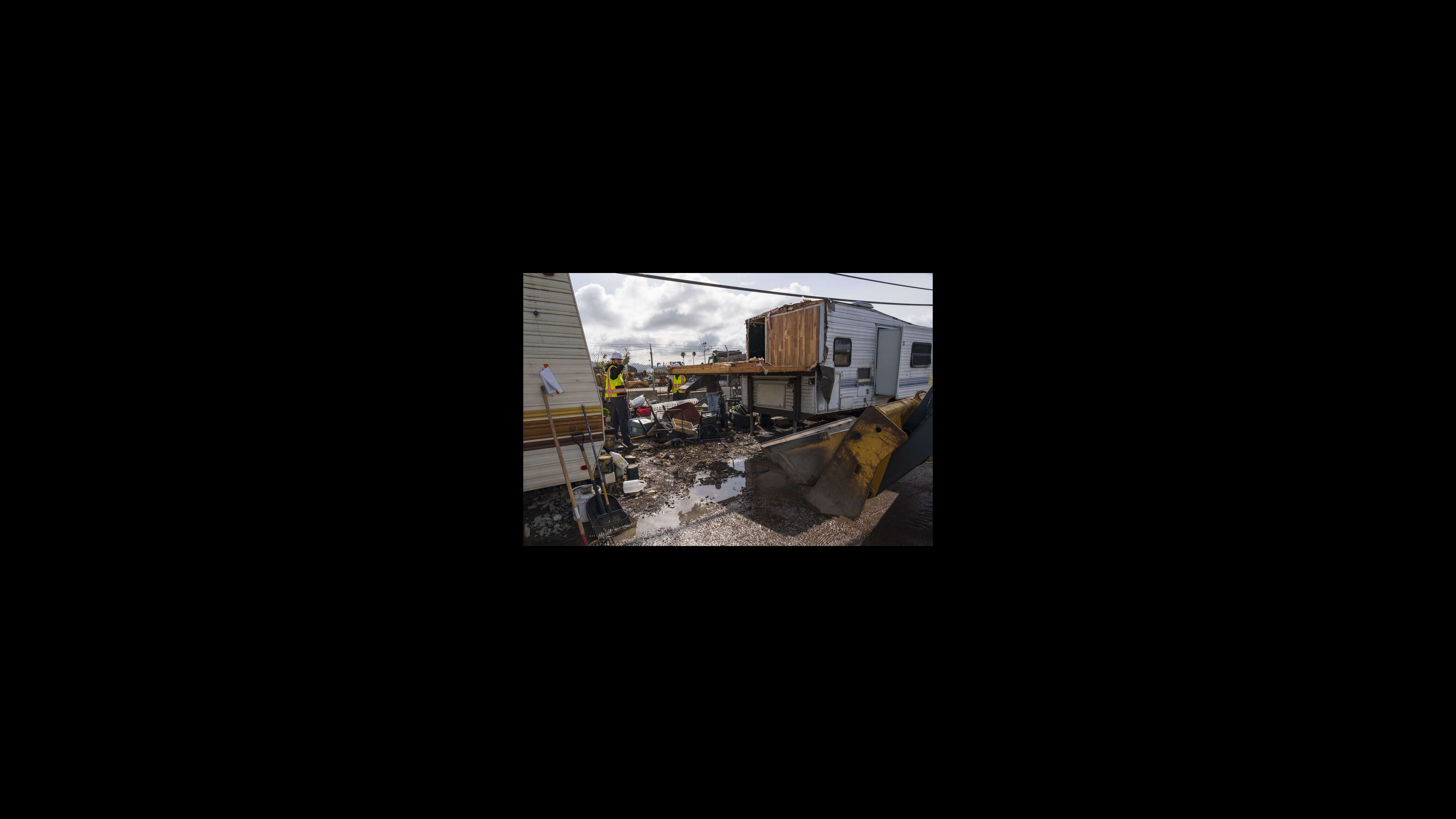 A Berkeley Public Works employee signals to the bulldozer driver to approach and pick up remaining debris after demolishing a second-story room extending from a trailer. The city strictly prohibits the construction of makeshift structures, including those attached to vehicles. A city worker motions to a bulldozer driver, directing him toward a wooden structure that was built as an addition on an RV. They are in the process of dismantling and removing the wooden structure. The bulldozer's scoop is visible in the right foreground.