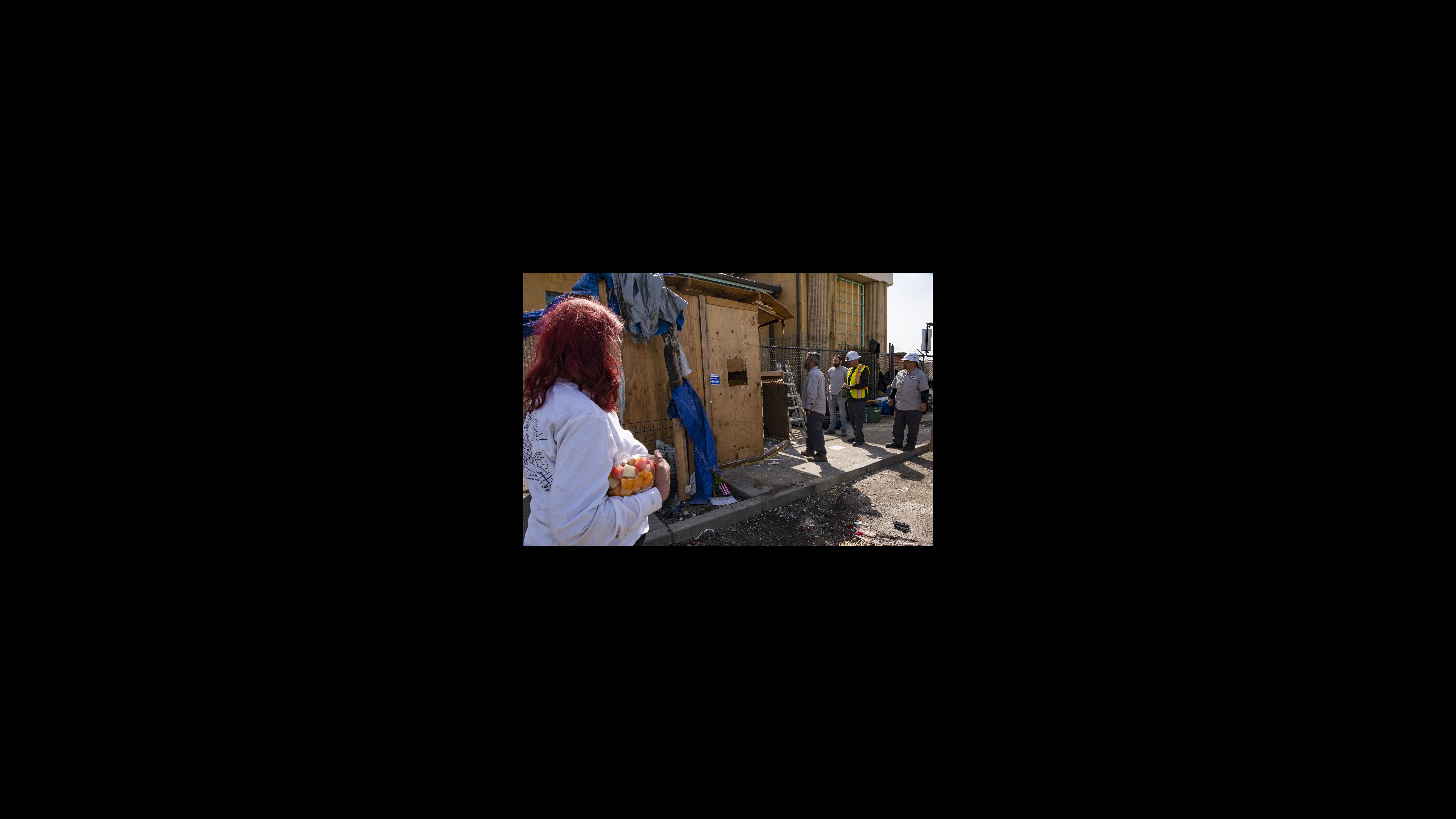 A Second Street resident watches city staffers prepare to demolish her home. Her son built the wooden structure, which protected her from the harsh atmospheric river storms last winter. Four city workers stand near a makeshift wooden structure that they are preparing to dismantle. A woman with red hair wearing a long-sleeved white T-shirt and holding a plastic container of cut fruit pieces stands in the foreground watching them.