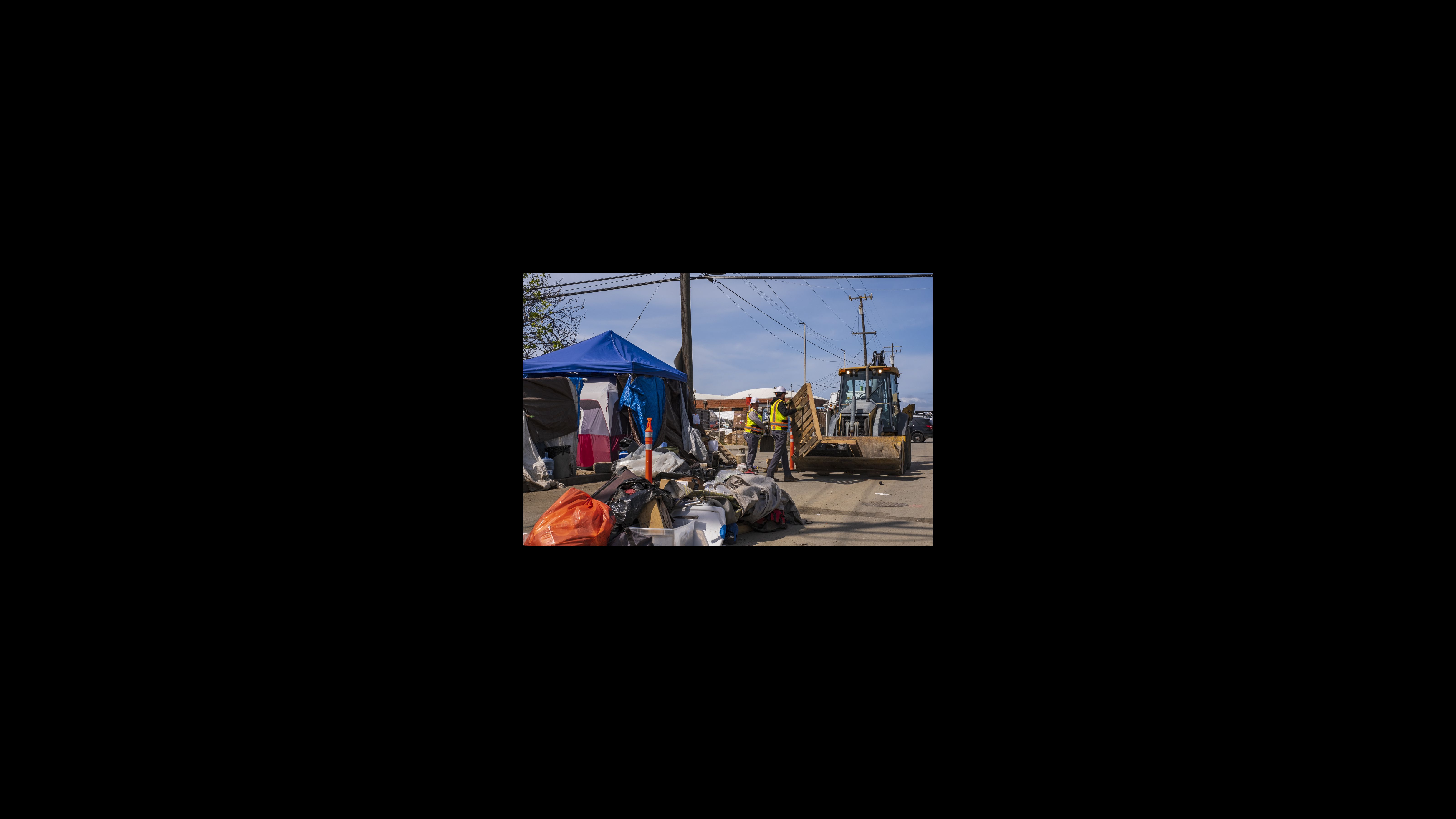 Second Street residents pile trash in the middle of the road for Public Works to pick up during a deep cleaning on May 9. The city said it would not enter anyone’s tents if residents participated in the cleanup and gave up bulky items voluntarily. Two city workers place wooden pallets for a bulldozer to carry away. They are in an area where items are being sorted into piles for storage and for discarding. There are tents on the left side of the image.
