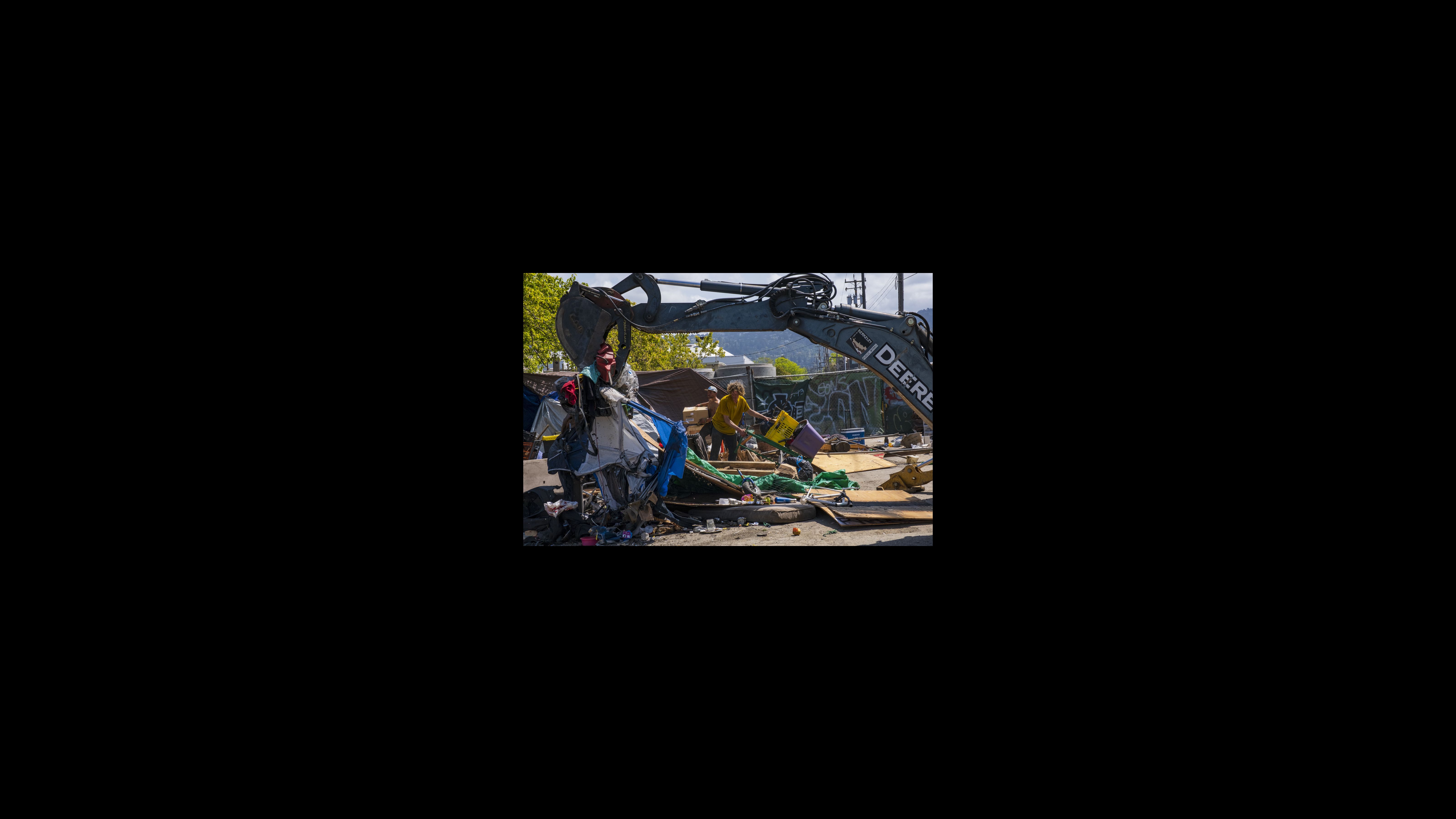 While a backhoe picks up a tent, Matty Henderson assists his neighbor Marjo Hughes in moving his personal belongings away from the green fence, clearing a path for incoming construction traffic. A backhoe picks up a tent and other discarded items to be thrown away. Two people in the background sort items to be kept or discarded.